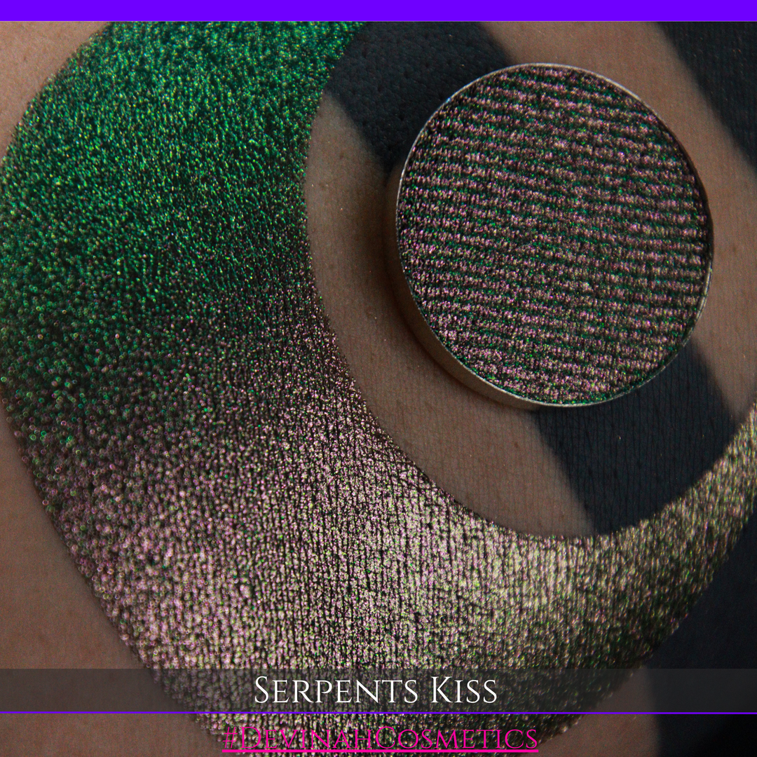 A round eyeshadow pan labeled 'SERPENTS KISS' made by Devinah Cosmetics, placed over a large crescent moon swatch of the same shimmering shifty eyeshadow on skin. The eyeshadow has a duochrome multichrome shift between colors greeen, olive brown, red.