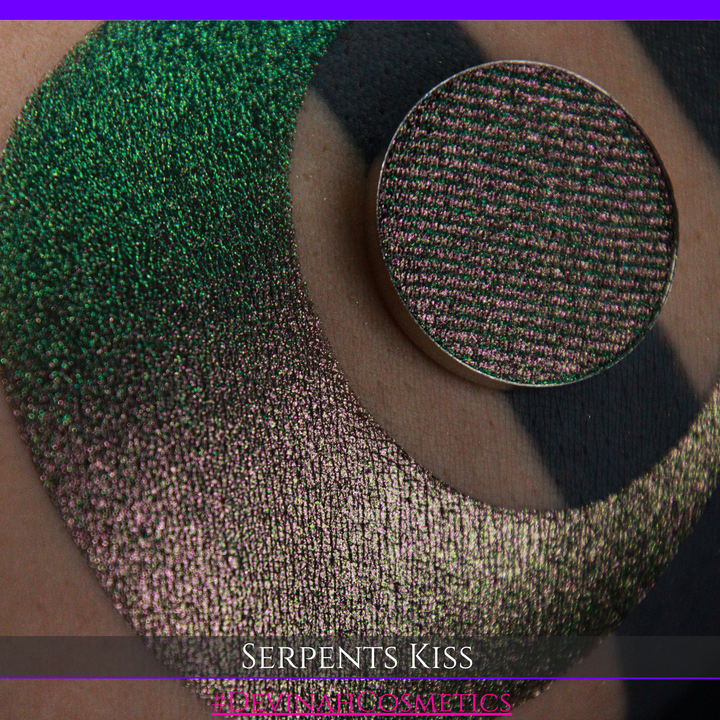 A round eyeshadow pan labeled 'SERPENTS KISS' made by Devinah Cosmetics, placed over a large crescent moon swatch of the same shimmering shifty eyeshadow on skin. The eyeshadow has a duochrome multichrome shift between colors greeen, olive brown, red.