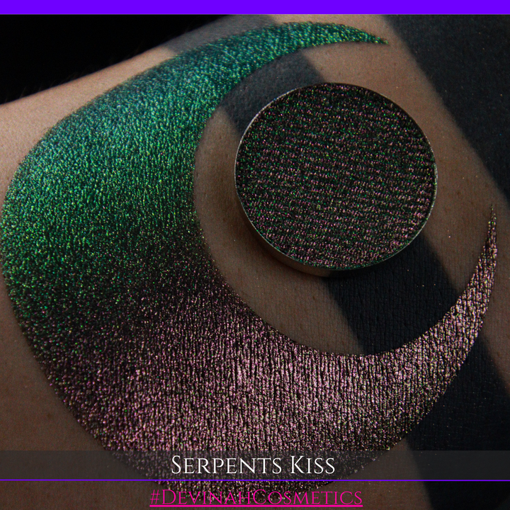A round eyeshadow pan labeled 'SERPENTS KISS' made by Devinah Cosmetics, placed over a large crescent moon swatch of the same shimmering shifty eyeshadow on skin. The eyeshadow has a duochrome multichrome shift between colors greeen, olive brown, red.