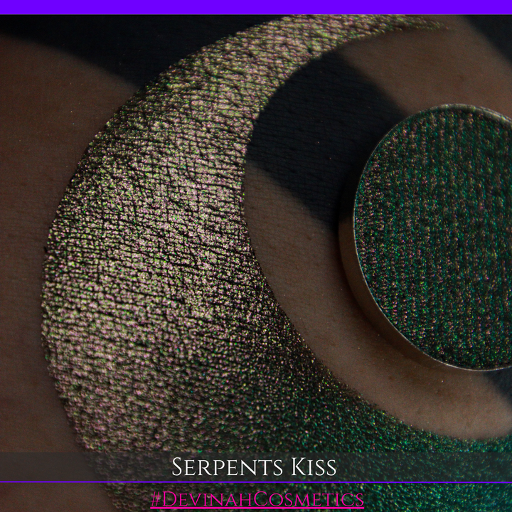 A round eyeshadow pan labeled 'SERPENTS KISS' made by Devinah Cosmetics, placed over a large crescent moon swatch of the same shimmering shifty eyeshadow on skin. The eyeshadow has a duochrome multichrome shift between colors greeen, olive brown, red.