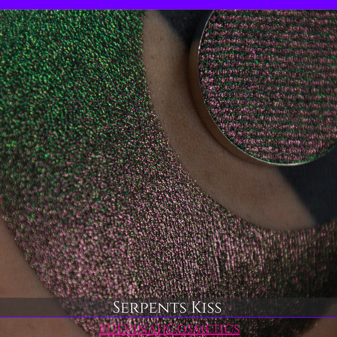 A round eyeshadow pan labeled 'SERPENTS KISS' made by Devinah Cosmetics, placed over a large crescent moon swatch of the same shimmering shifty eyeshadow on skin. The eyeshadow has a duochrome multichrome shift between colors greeen, olive brown, red.