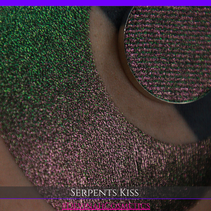 A round eyeshadow pan labeled 'SERPENTS KISS' made by Devinah Cosmetics, placed over a large crescent moon swatch of the same shimmering shifty eyeshadow on skin. The eyeshadow has a duochrome multichrome shift between colors greeen, olive brown, red.