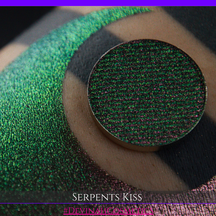 A round eyeshadow pan labeled 'SERPENTS KISS' made by Devinah Cosmetics, placed over a large crescent moon swatch of the same shimmering shifty eyeshadow on skin. The eyeshadow has a duochrome multichrome shift between colors greeen, olive brown, red.