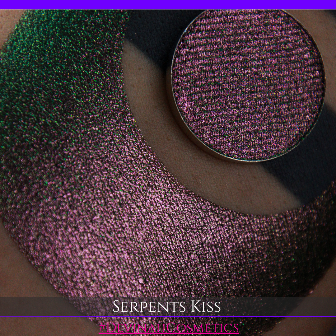 A round eyeshadow pan labeled 'SERPENTS KISS' made by Devinah Cosmetics, placed over a large crescent moon swatch of the same shimmering shifty eyeshadow on skin. The eyeshadow has a duochrome multichrome shift between colors greeen, olive brown, red.