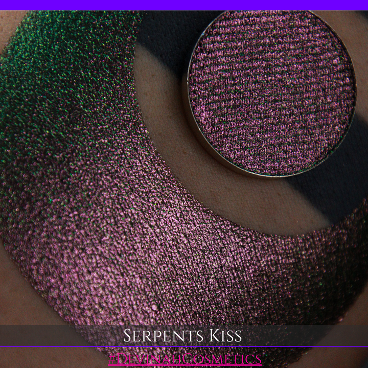 A round eyeshadow pan labeled 'SERPENTS KISS' made by Devinah Cosmetics, placed over a large crescent moon swatch of the same shimmering shifty eyeshadow on skin. The eyeshadow has a duochrome multichrome shift between colors greeen, olive brown, red.