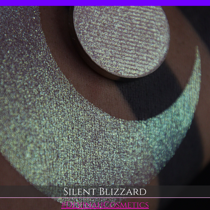 A close-up of a person's arm with a large eyeshadow swatch in a crescent shape and a matching round makeup pan labeled 'Silent Blizzard' hand made by Devinah Cosmetics. The product color shifts between silver, green, and gold depending on the angle, with a dark background.