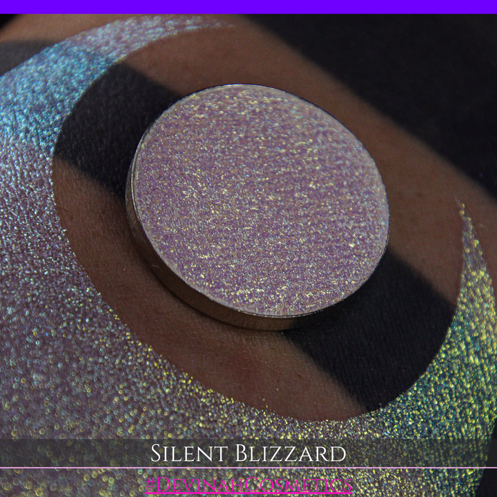 A close-up of a person's arm with a large eyeshadow swatch in a crescent shape and a matching round makeup pan labeled 'Silent Blizzard' hand made by Devinah Cosmetics. The product color shifts between silver, green, and gold depending on the angle, with a dark background.