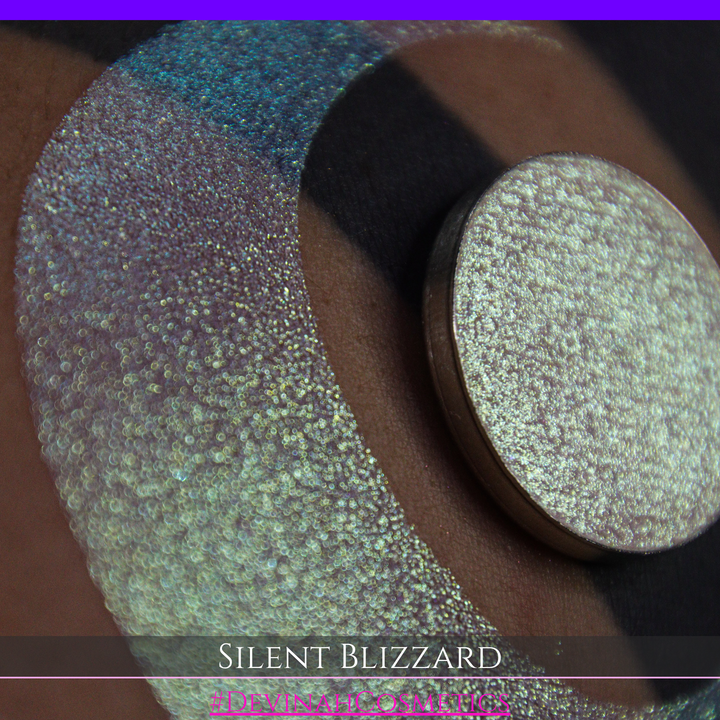 A close-up of a person's arm with a large eyeshadow swatch in a crescent shape and a matching round makeup pan labeled 'Silent Blizzard' hand made by Devinah Cosmetics. The product color shifts between silver, green, and gold depending on the angle, with a dark background.