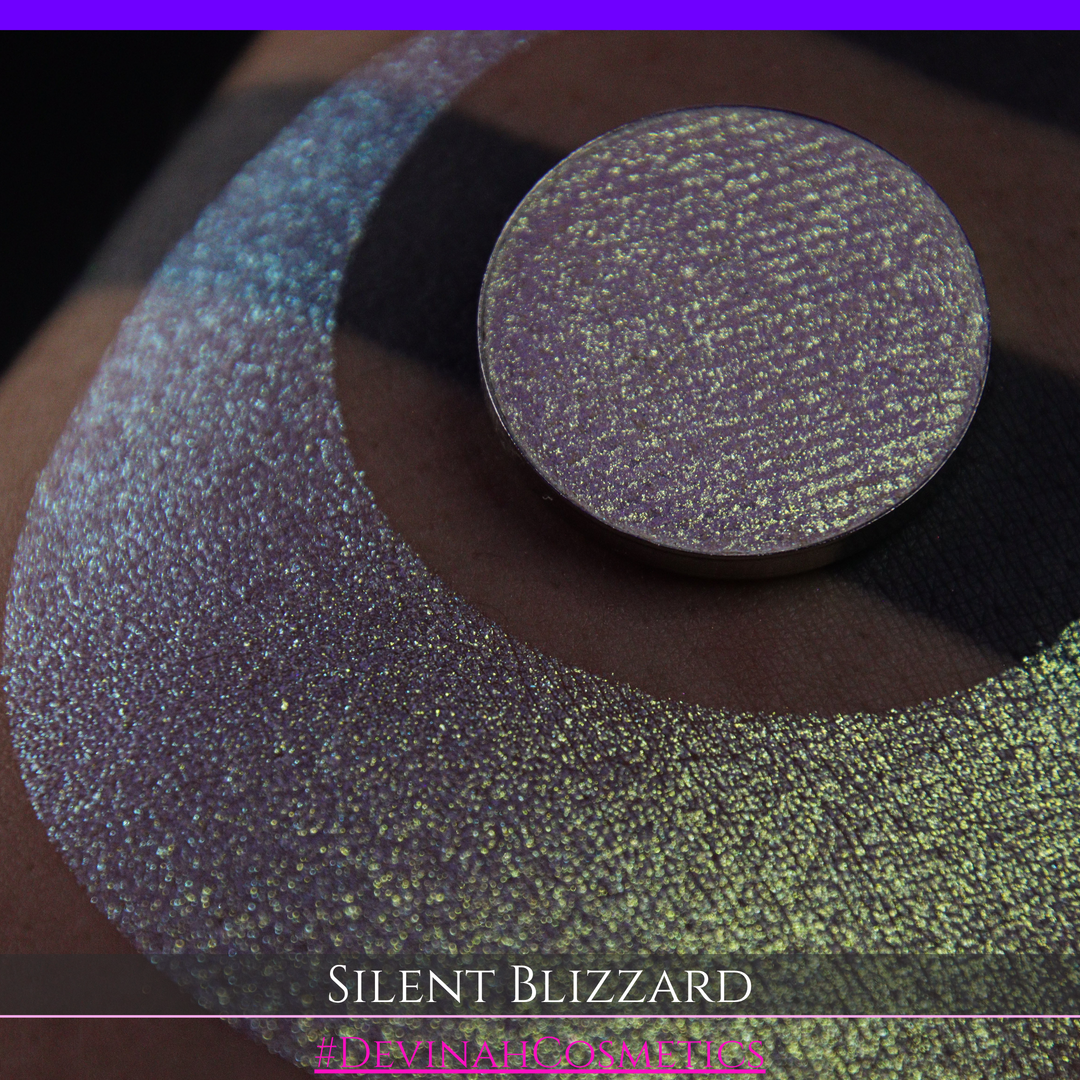 A close-up of a person's arm with a large eyeshadow swatch in a crescent shape and a matching round makeup pan labeled 'Silent Blizzard' hand made by Devinah Cosmetics. The product color shifts between silver, green, and gold depending on the angle, with a dark background.