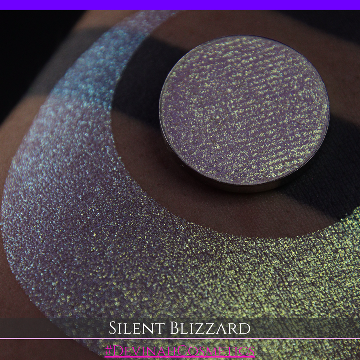 A close-up of a person's arm with a large eyeshadow swatch in a crescent shape and a matching round makeup pan labeled 'Silent Blizzard' hand made by Devinah Cosmetics. The product color shifts between silver, green, and gold depending on the angle, with a dark background.