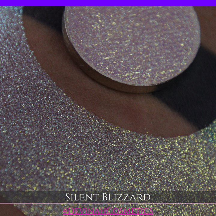 A close-up of a person's arm with a large eyeshadow swatch in a crescent shape and a matching round makeup pan labeled 'Silent Blizzard' hand made by Devinah Cosmetics. The product color shifts between silver, green, and gold depending on the angle, with a dark background.