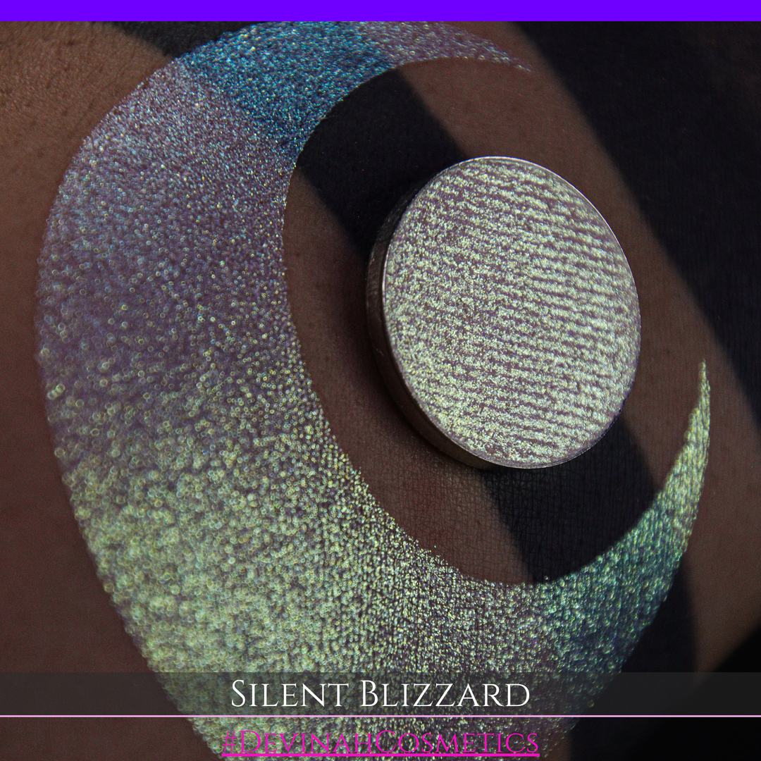 A close-up of a person's arm with a large eyeshadow swatch in a crescent shape and a matching round makeup pan labeled 'Silent Blizzard' hand made by Devinah Cosmetics. The product color shifts between silver, green, and gold depending on the angle, with a dark background.