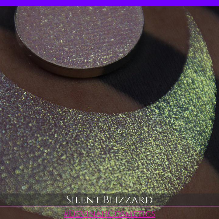 A close-up of a person's arm with a large eyeshadow swatch in a crescent shape and a matching round makeup pan labeled 'Silent Blizzard' hand made by Devinah Cosmetics. The product color shifts between silver, green, and gold depending on the angle, with a dark background.