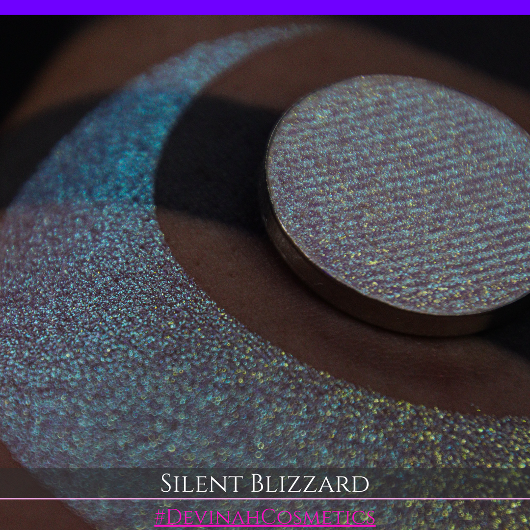 A close-up of a person's arm with a large eyeshadow swatch in a crescent shape and a matching round makeup pan labeled 'Silent Blizzard' hand made by Devinah Cosmetics. The product color shifts between silver, green, and gold depending on the angle, with a dark background.