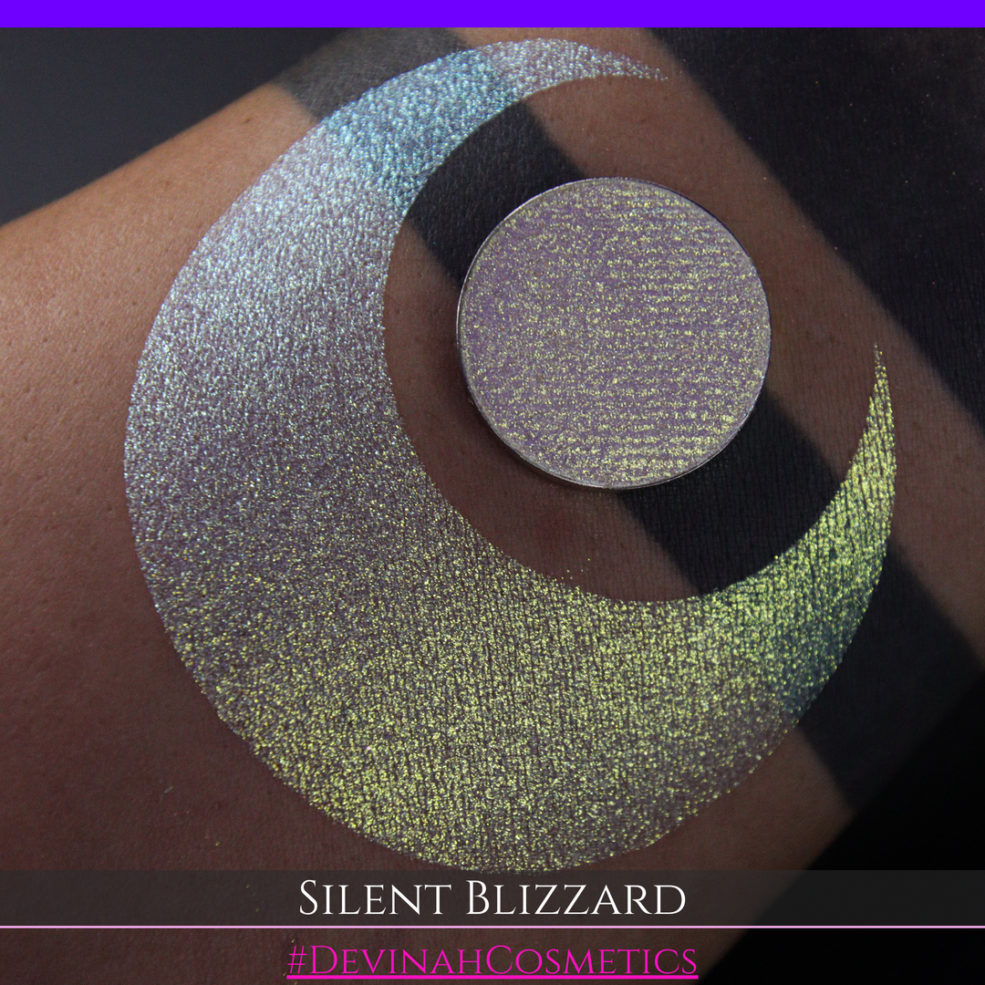 A close-up of a person's arm with a large eyeshadow swatch in a crescent shape and a matching round makeup pan labeled 'Silent Blizzard' hand made by Devinah Cosmetics. The product color shifts between silver, green, and gold depending on the angle, with a dark background.