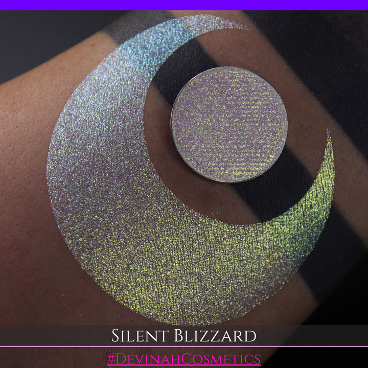 A close-up of a person's arm with a large eyeshadow swatch in a crescent shape and a matching round makeup pan labeled 'Silent Blizzard' hand made by Devinah Cosmetics. The product color shifts between silver, green, and gold depending on the angle, with a dark background.
