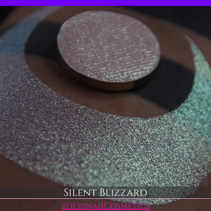 A close-up of a person's arm with a large eyeshadow swatch in a crescent shape and a matching round makeup pan labeled 'Silent Blizzard' hand made by Devinah Cosmetics. The product color shifts between silver, green, and gold depending on the angle, with a dark background.