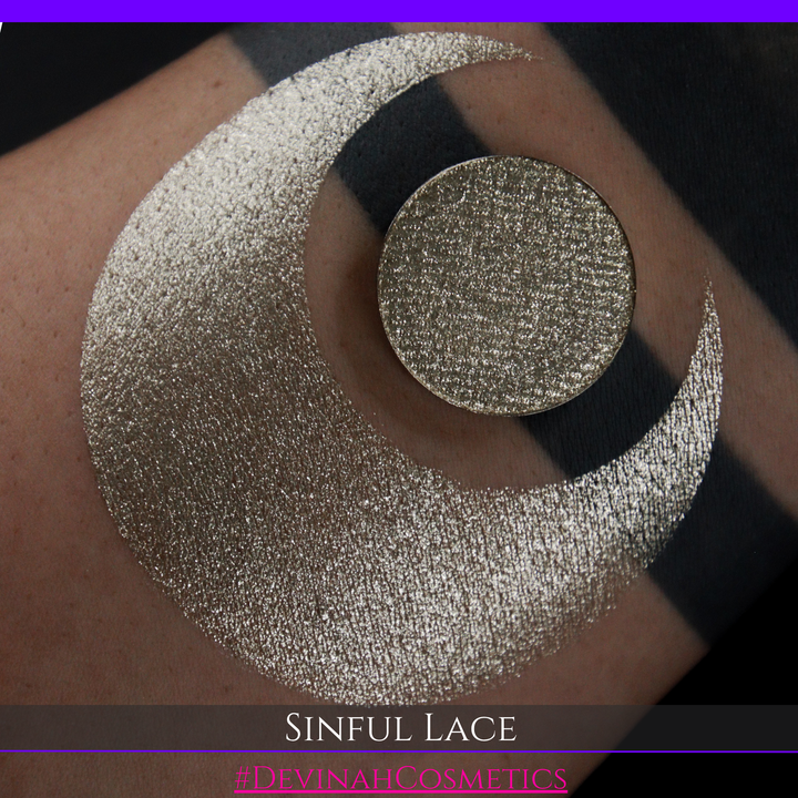 A round eyeshadow pan labeled 'SINFUL LACE' made by Devinah Cosmetics, placed over a large crescent moon swatch of the same shimmering shifty eyeshadow on skin. The eyeshadow has colors of sparkly beige creme tan shimmer.