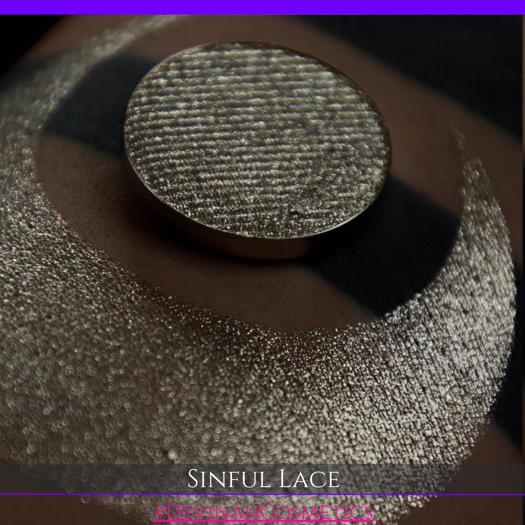 A round eyeshadow pan labeled 'SINFUL LACE' made by Devinah Cosmetics, placed over a large crescent moon swatch of the same shimmering shifty eyeshadow on skin. The eyeshadow has colors of sparkly beige creme tan shimmer.