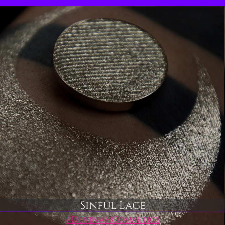 A round eyeshadow pan labeled 'SINFUL LACE' made by Devinah Cosmetics, placed over a large crescent moon swatch of the same shimmering shifty eyeshadow on skin. The eyeshadow has colors of sparkly beige creme tan shimmer.