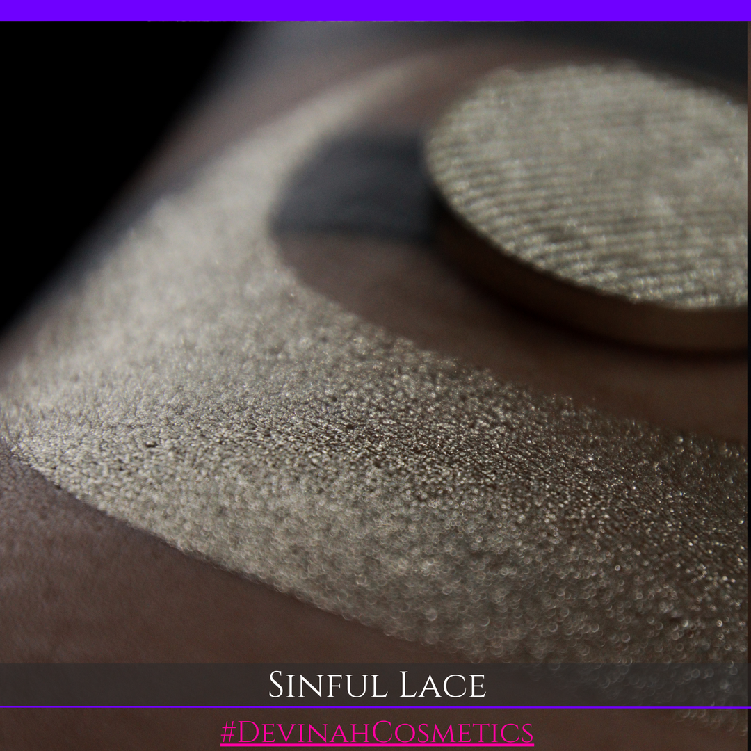 A round eyeshadow pan labeled 'SINFUL LACE' made by Devinah Cosmetics, placed over a large crescent moon swatch of the same shimmering shifty eyeshadow on skin. The eyeshadow has colors of sparkly beige creme tan shimmer.