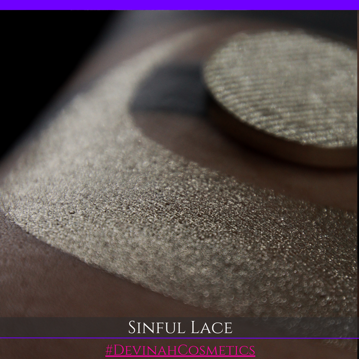 A round eyeshadow pan labeled 'SINFUL LACE' made by Devinah Cosmetics, placed over a large crescent moon swatch of the same shimmering shifty eyeshadow on skin. The eyeshadow has colors of sparkly beige creme tan shimmer.