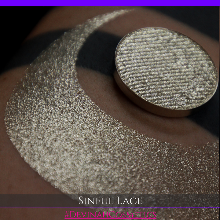 A round eyeshadow pan labeled 'SINFUL LACE' made by Devinah Cosmetics, placed over a large crescent moon swatch of the same shimmering shifty eyeshadow on skin. The eyeshadow has colors of sparkly beige creme tan shimmer.