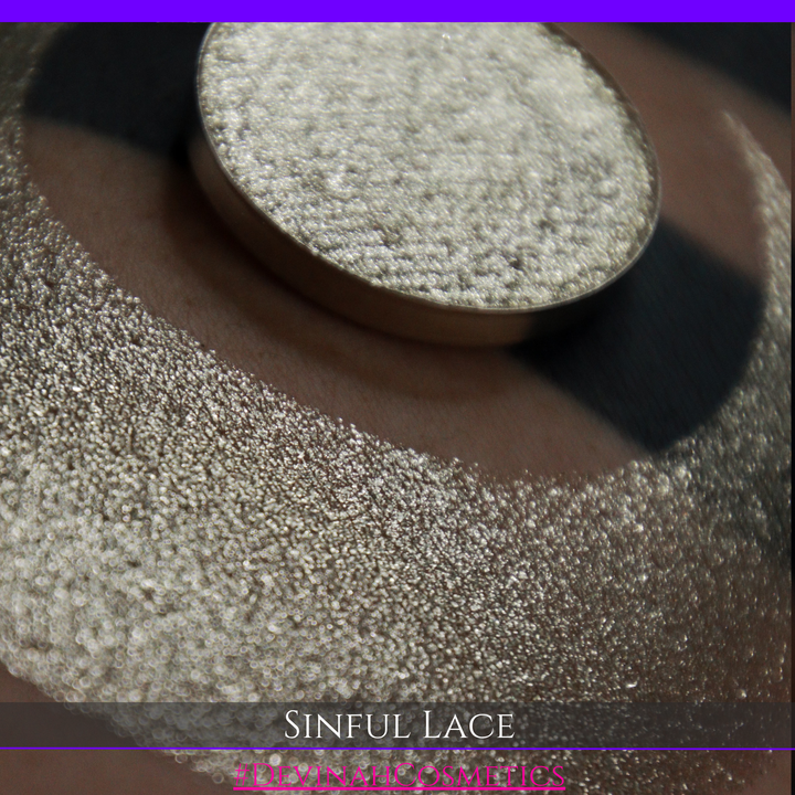 A round eyeshadow pan labeled 'SINFUL LACE' made by Devinah Cosmetics, placed over a large crescent moon swatch of the same shimmering shifty eyeshadow on skin. The eyeshadow has colors of sparkly beige creme tan shimmer.