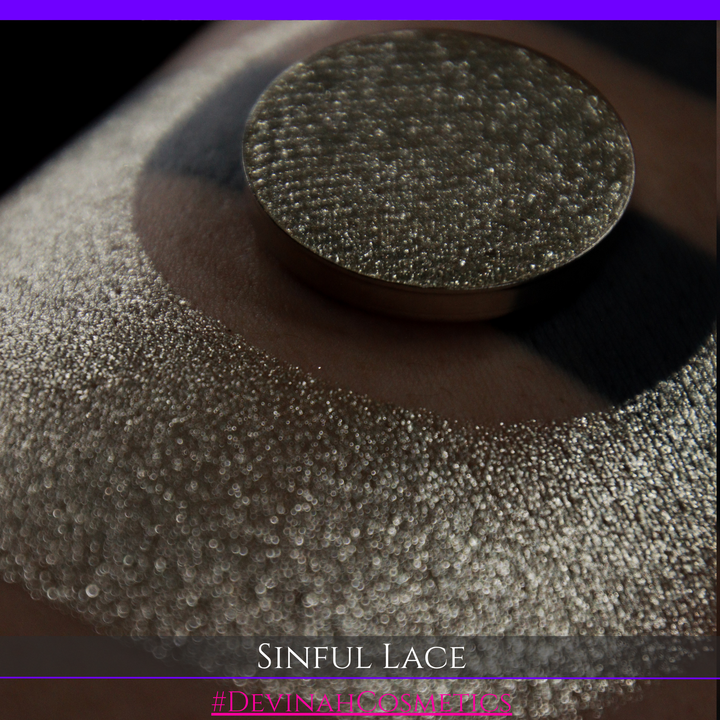 A round eyeshadow pan labeled 'SINFUL LACE' made by Devinah Cosmetics, placed over a large crescent moon swatch of the same shimmering shifty eyeshadow on skin. The eyeshadow has colors of sparkly beige creme tan shimmer.