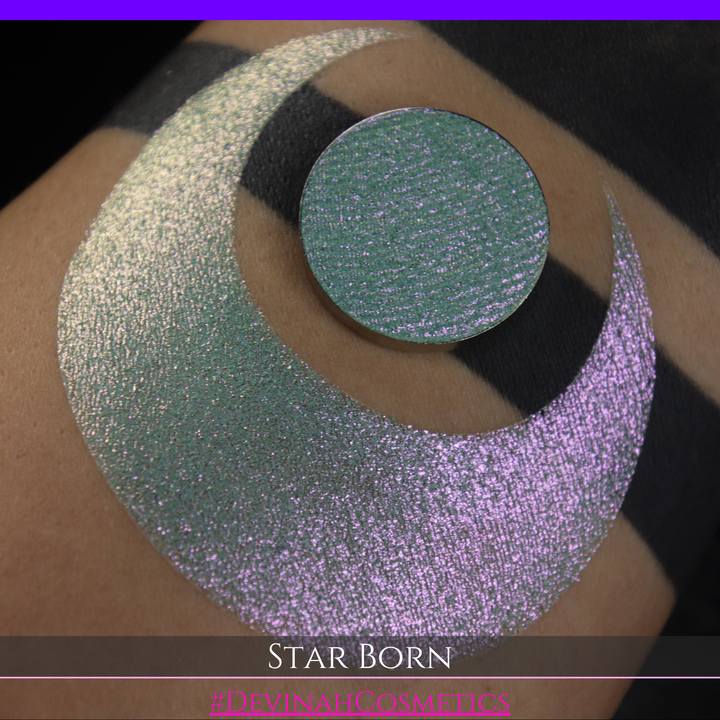 A close-up of a person's skin with a large iridescent crescent moon makeup swatch and a matching circular eyeshadow pan in the center. The text 'STAR BORN' and 'Devinah Cosmetics' appears at the bottom.
