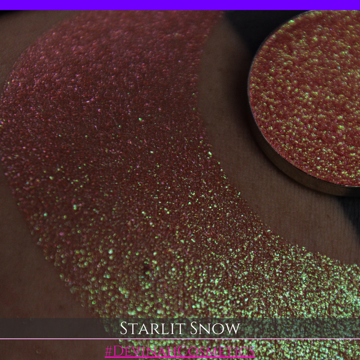 A close-up of a person's arm with a large eyeshadow swatch in a crescent shape and a matching round eyeshadow pan placed in the center. Text at the bottom reads 'STARLIT SNOW' and '#DEVINAHCosmetics'.