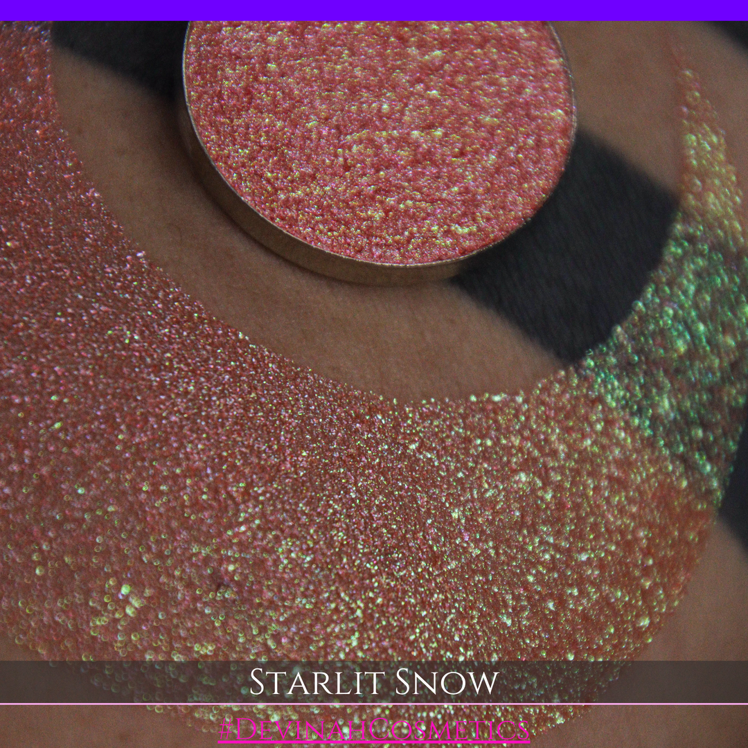 A close-up of a person's arm with a large eyeshadow swatch in a crescent shape and a matching round eyeshadow pan placed in the center. Text at the bottom reads 'STARLIT SNOW' and '#DEVINAHCosmetics'.