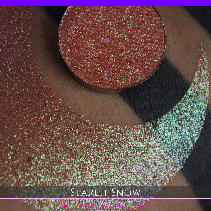 A close-up of a person's arm with a large eyeshadow swatch in a crescent shape and a matching round eyeshadow pan placed in the center. Text at the bottom reads 'STARLIT SNOW' and '#DEVINAHCosmetics'.