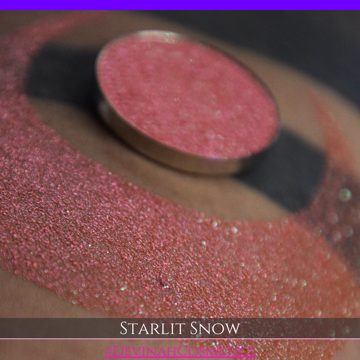 A close-up of a person's arm with a large eyeshadow swatch in a crescent shape and a matching round eyeshadow pan placed in the center. Text at the bottom reads 'STARLIT SNOW' and '#DEVINAHCosmetics'.