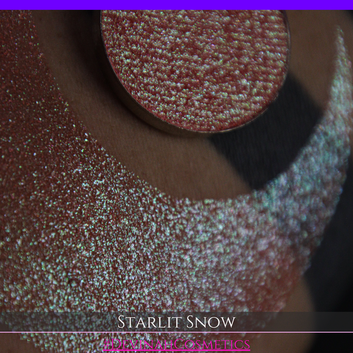 A close-up of a person's arm with a large eyeshadow swatch in a crescent shape and a matching round eyeshadow pan placed in the center. Text at the bottom reads 'STARLIT SNOW' and '#DEVINAHCosmetics'.