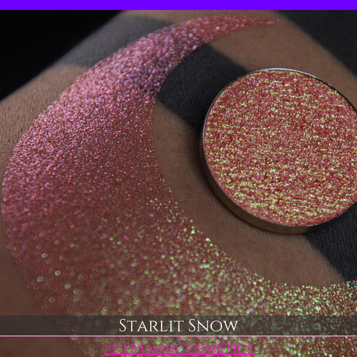A close-up of a person's arm with a large eyeshadow swatch in a crescent shape and a matching round eyeshadow pan placed in the center. Text at the bottom reads 'STARLIT SNOW' and '#DEVINAHCosmetics'.