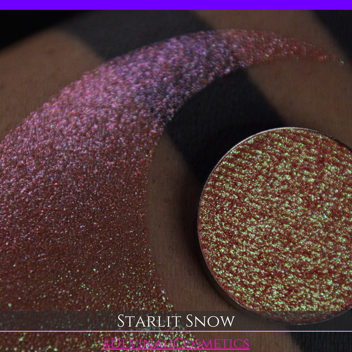 A close-up of a person's arm with a large eyeshadow swatch in a crescent shape and a matching round eyeshadow pan placed in the center. Text at the bottom reads 'STARLIT SNOW' and '#DEVINAHCosmetics'.