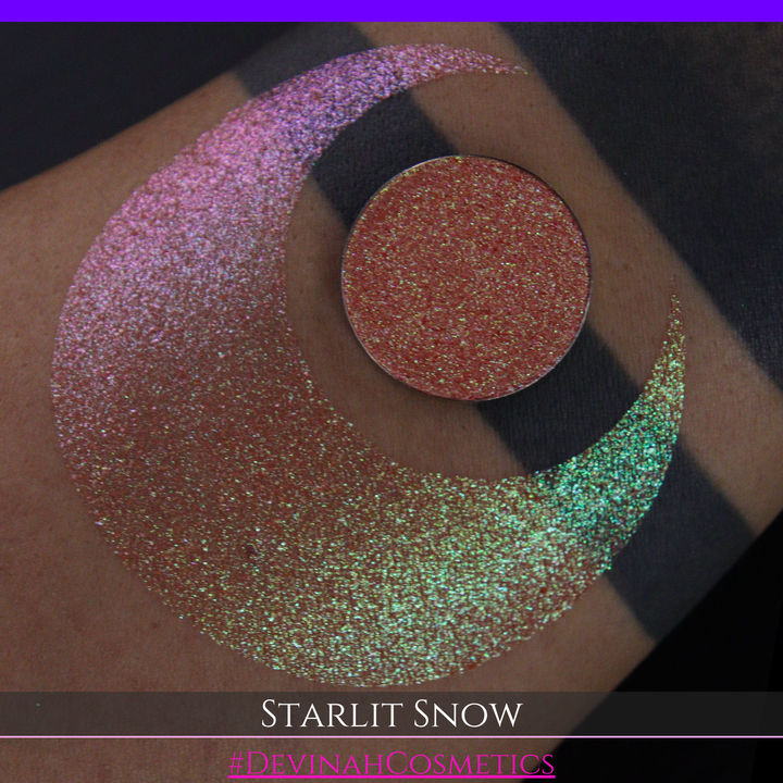 A close-up of a person's arm with a large eyeshadow swatch in a crescent shape and a matching round eyeshadow pan placed in the center. Text at the bottom reads 'STARLIT SNOW' and '#DEVINAHCosmetics'.