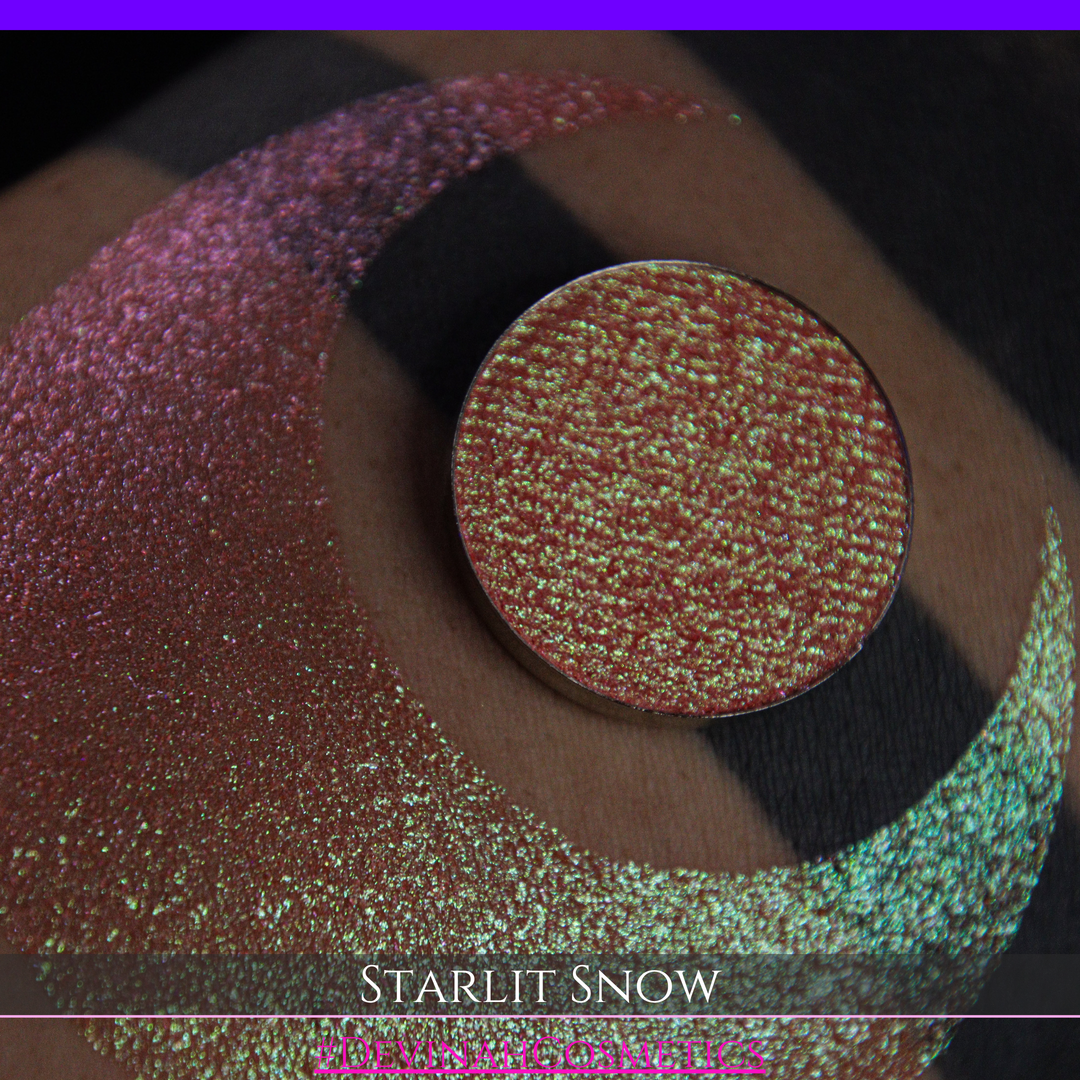 A close-up of a person's arm with a large eyeshadow swatch in a crescent shape and a matching round eyeshadow pan placed in the center. Text at the bottom reads 'STARLIT SNOW' and '#DEVINAHCosmetics'.