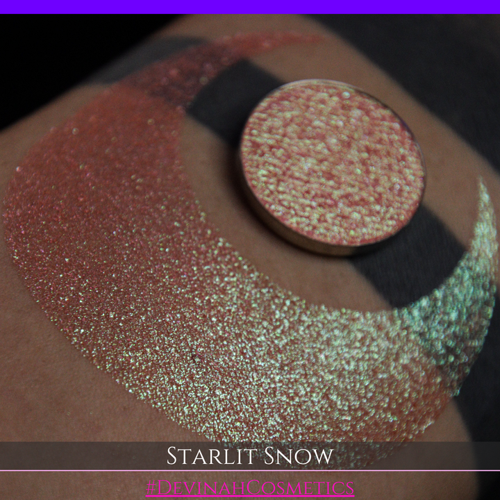 A close-up of a person's arm with a large eyeshadow swatch in a crescent shape and a matching round eyeshadow pan placed in the center. Text at the bottom reads 'STARLIT SNOW' and '#DEVINAHCosmetics'.