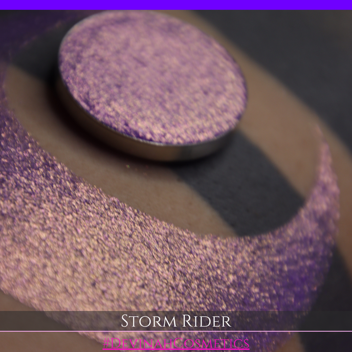 A close-up of a person's arm with a large swatch of glittery purple eyeshadow and a matching round pan of the same shade, labeled 'STORM RIDER' with '#DEVINAH COSMETICS'.