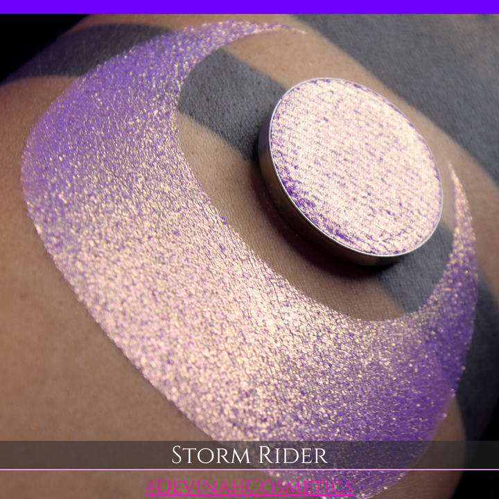 A close-up of a person's arm with a large swatch of glittery purple eyeshadow and a matching round pan of the same shade, labeled 'STORM RIDER' with '#DEVINAH COSMETICS'.