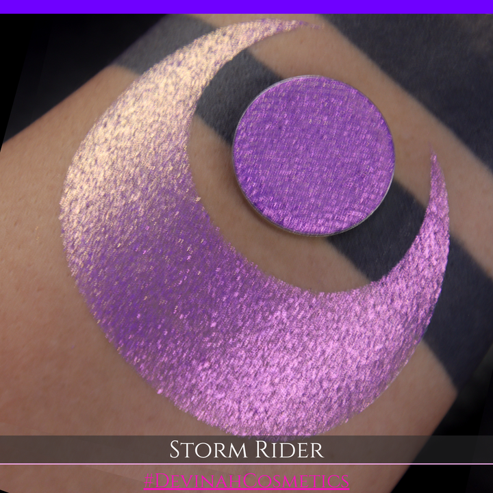 A close-up of a person's arm with a large swatch of glittery purple eyeshadow and a matching round pan of the same shade, labeled 'STORM RIDER' with '#DEVINAH COSMETICS'.