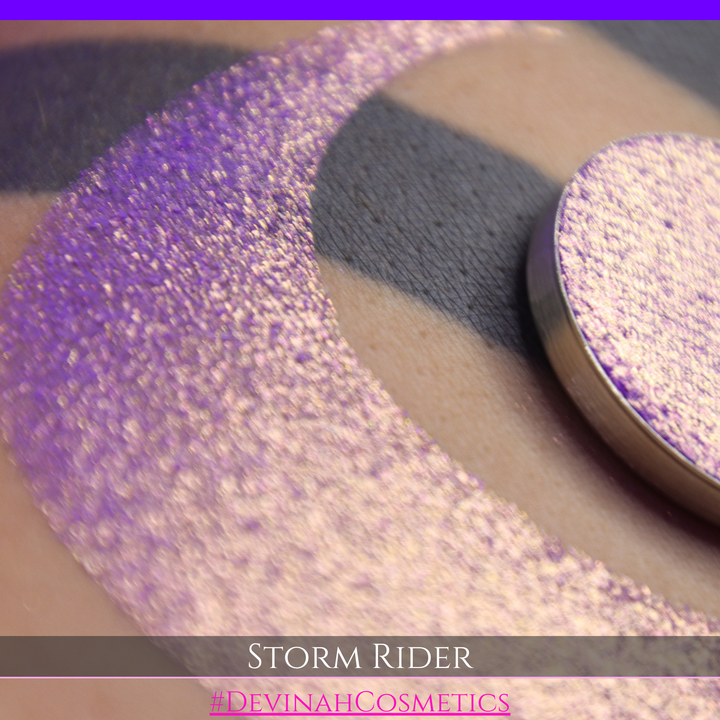 A close-up of a person's arm with a large swatch of glittery purple eyeshadow and a matching round pan of the same shade, labeled 'STORM RIDER' with '#DEVINAH COSMETICS'.