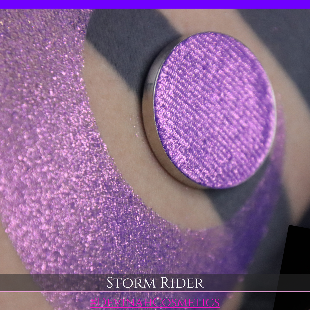 A close-up of a person's arm with a large swatch of glittery purple eyeshadow and a matching round pan of the same shade, labeled 'STORM RIDER' with '#DEVINAH COSMETICS'.