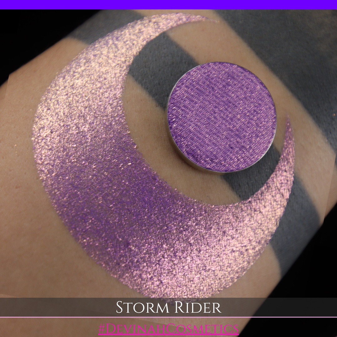 A close-up of a person's arm with a large swatch of glittery purple eyeshadow and a matching round pan of the same shade, labeled 'STORM RIDER' with '#DEVINAH COSMETICS'.