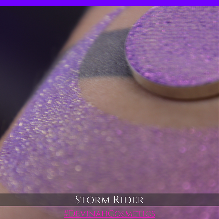 A close-up of a person's arm with a large swatch of glittery purple eyeshadow and a matching round pan of the same shade, labeled 'STORM RIDER' with '#DEVINAH COSMETICS'.