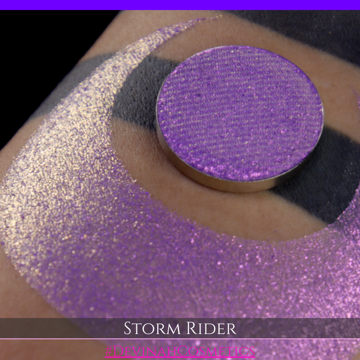 A close-up of a person's arm with a large swatch of glittery purple eyeshadow and a matching round pan of the same shade, labeled 'STORM RIDER' with '#DEVINAH COSMETICS'.
