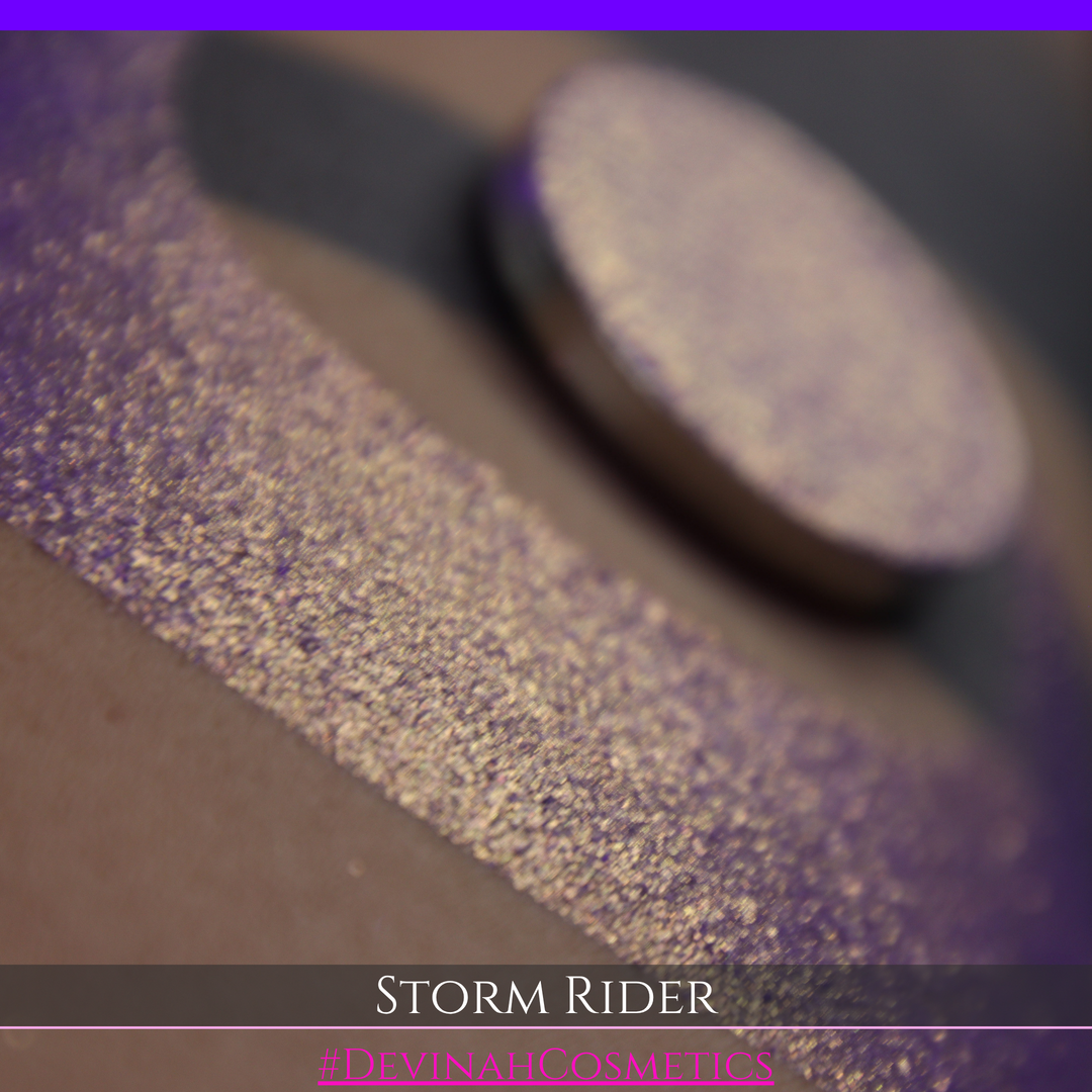 A close-up of a person's arm with a large swatch of glittery purple eyeshadow and a matching round pan of the same shade, labeled 'STORM RIDER' with '#DEVINAH COSMETICS'.