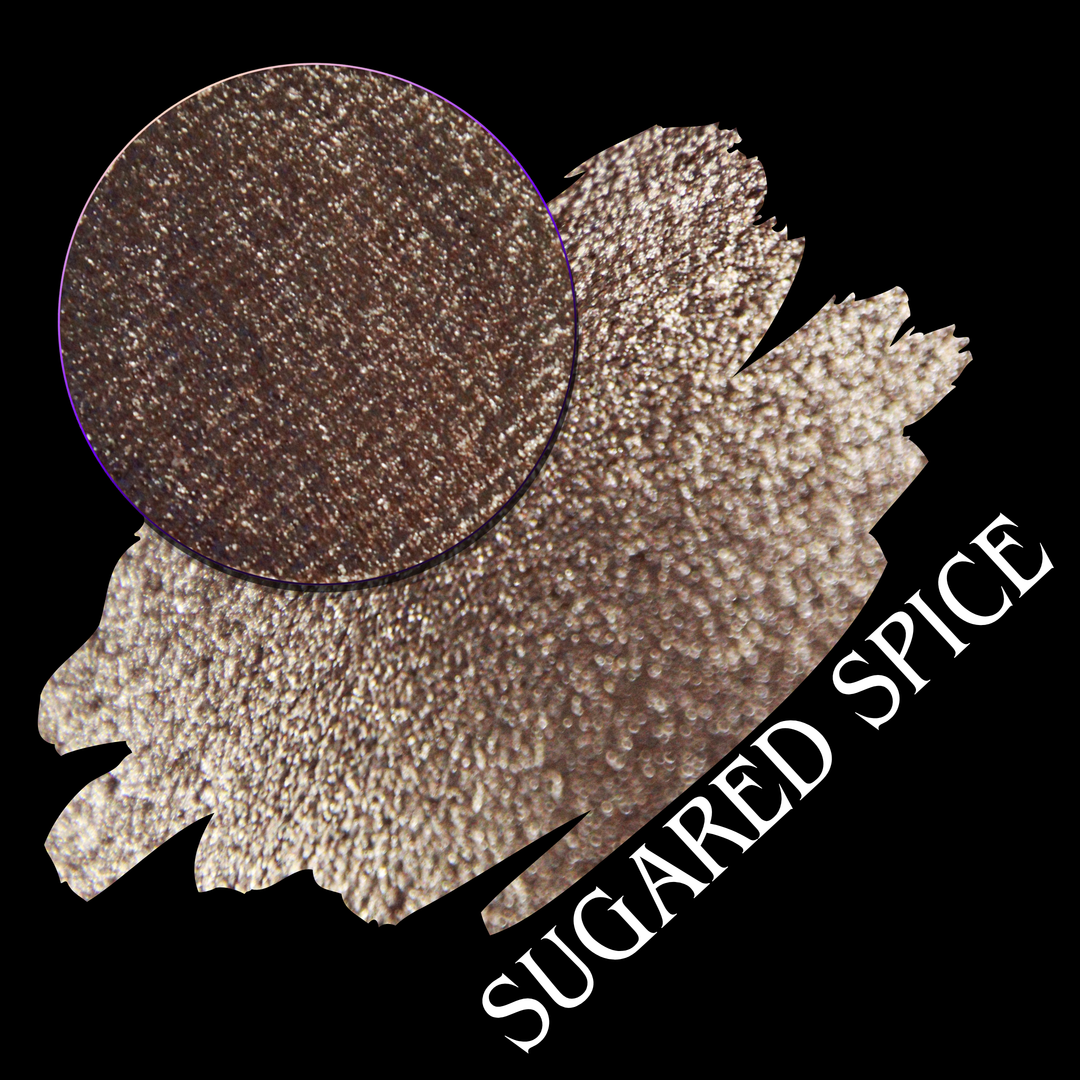 A round eyeshadow pan labeled 'SUGARED SPICE' made by Devinah Cosmetics, placed over a large crescent moon swatch of the same shimmering shifty eyeshadow on skin. The eyeshadow has sparkly colors of beige creme shimmer.